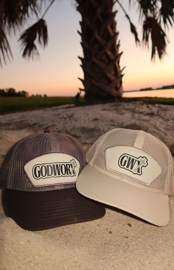 Two caps with godworx branding on a sandy surface with a sunset and palm tree in the background.