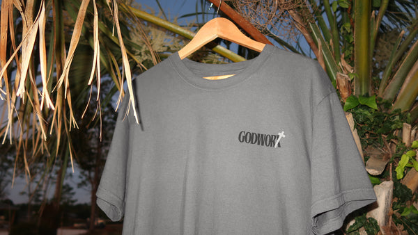 Gray t-shirt with 'GODWORK' logo on a hanger against a natural background