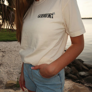 Person wearing a ivory colored t-shirt with 'GODWORX' logo by a body of water.
